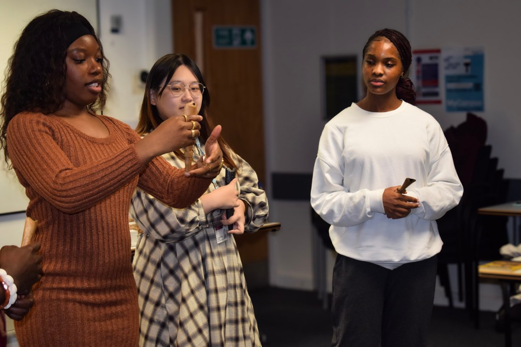 A group of participants engaged in an Igbo Flute Workshop at University College London, with one person demonstrating how to hold a flute, while others observe.